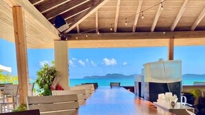 A long wooden table with benches, a large window with a view of the sea, and a ceiling with wooden beams and string lights.