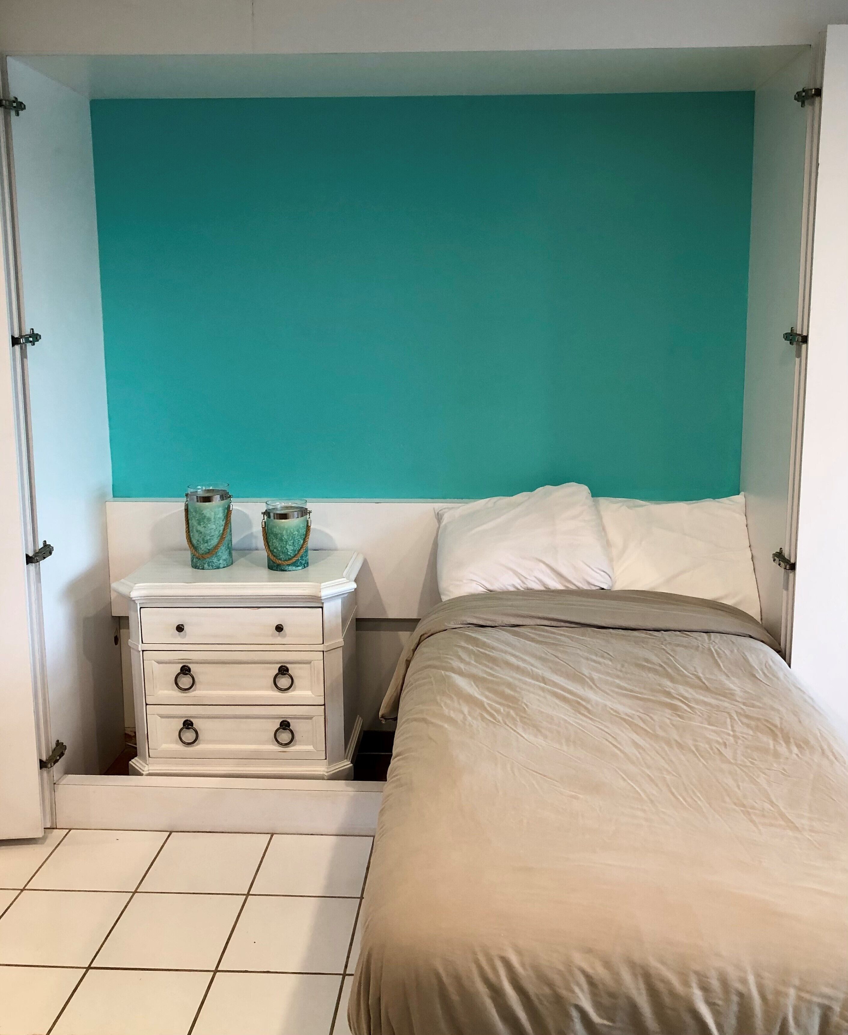 A bedroom with a teal accent wall, a white bedside table with drawers, a bed with a beige comforter, and two decorative glass vases on the table.