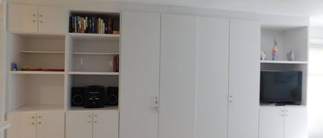 Una habitación amplia con armarios blancos, un televisor de pantalla plana y una estantería.