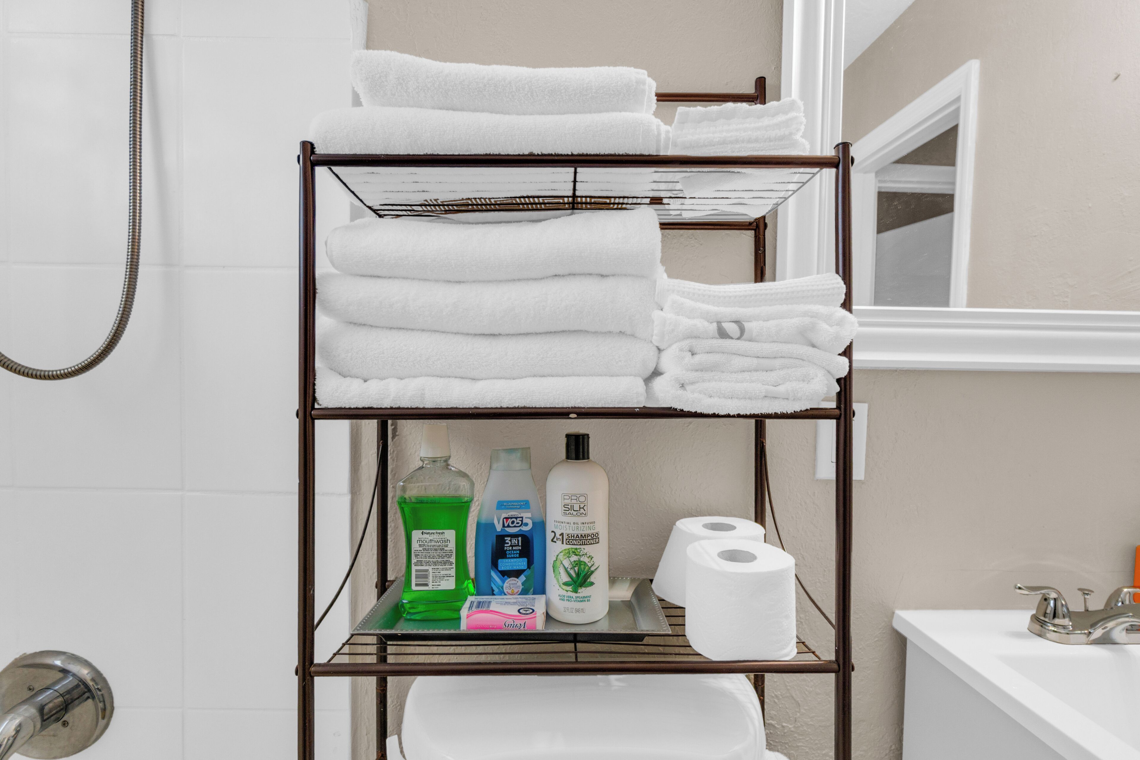 Une salle de bain avec une étagère en métal où sont posés des serviettes blanches, des articles de toilette et un rouleau de papier toilette.