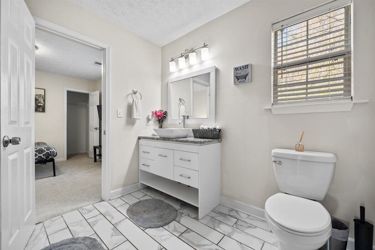 A bathroom with a white vanity, a large mirror, a toilet, and a window with blinds.