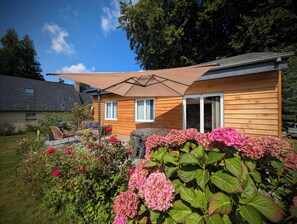 Ein Holzhaus mit Terrasse, überdachter Sitzgelegenheit und pinkfarbenen Hortensien im Garten.