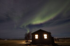 Une petite cabane en bois avec deux fenêtres, située dans une zone herbeuse, d’où l’on peut admirer le ciel nocturne.
