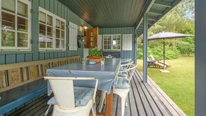 Ett takförsett utomhusområde med ett bord och stolar, ett trädäck och utsikt över den omgivande vegetationen.