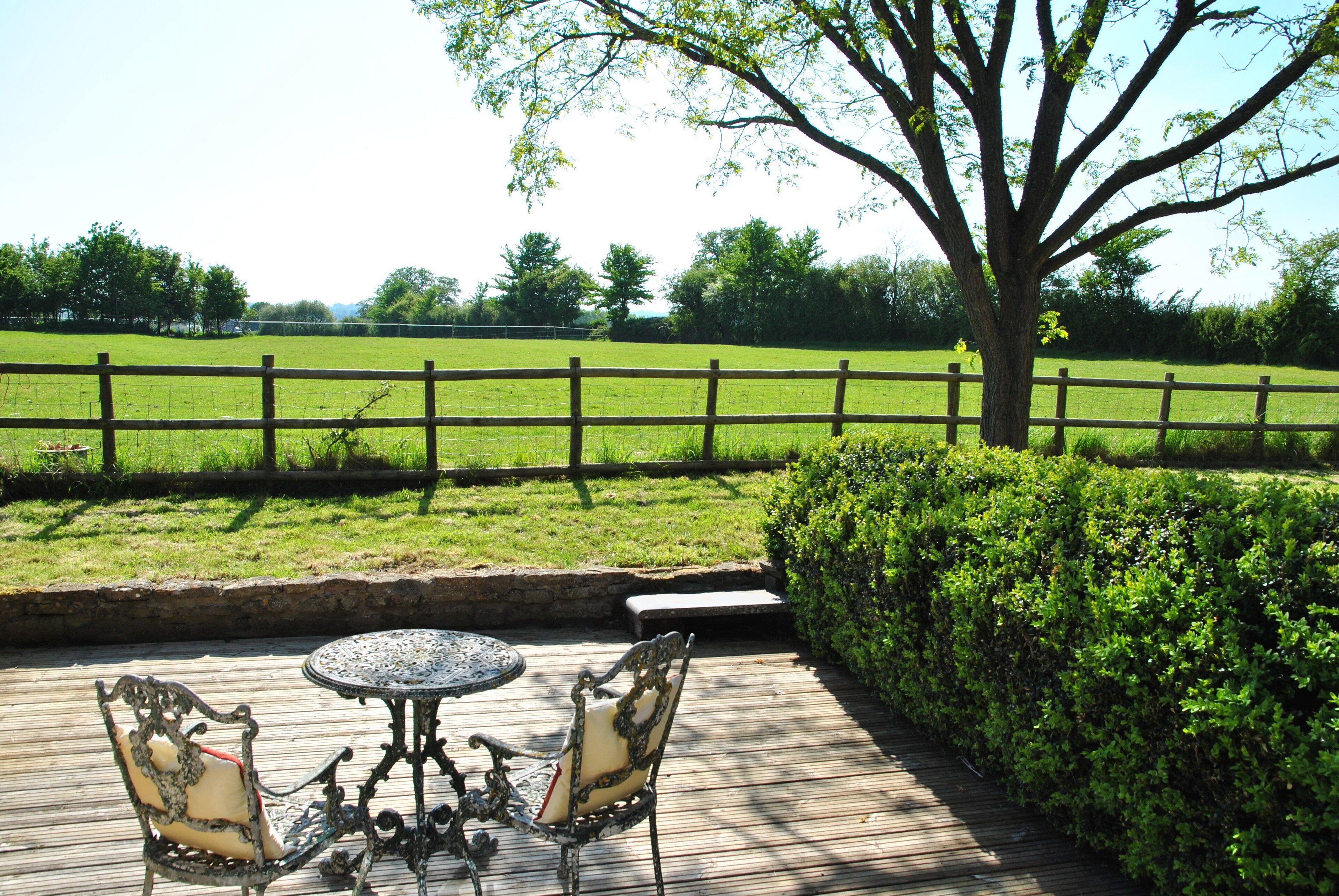Une terrasse extérieure avec une table et des chaises, une plateforme en bois et une vue sur un champ verdoyant et une clôture.