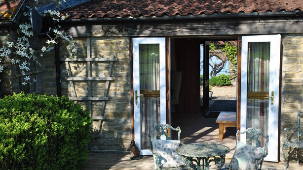 Un bâtiment en pierre doté d’une terrasse en bois, de deux tables avec des chaises et d’un banc.