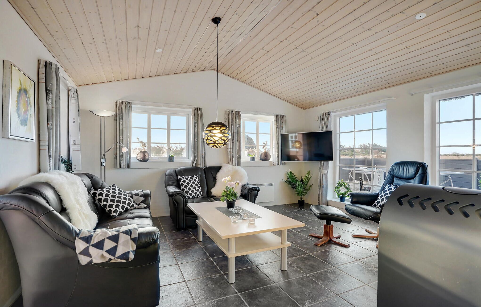 A modern living room with a wooden ceiling, large windows, a flat-screen TV, and leather furniture.