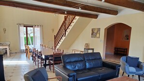 Ein großzügiger Wohnbereich mit einer großen Ledersofa, einem Holztisch und gemusterten Stühlen. Ein Esstisch mit Stühlen befindet sich in der Nähe eines Fensters mit Vorhängen. Eine Treppe mit Holzbalkonen führt auf eine obere Ebene.
