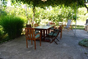 An outdoor patio with a wooden table and chairs, surrounded by greenery and a concrete floor. - Rustic House Miketa - Family Summertime (Preko)