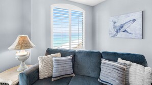 A corner room with a blue sofa, a lamp, a window with blinds, and a whale painting on the wall.