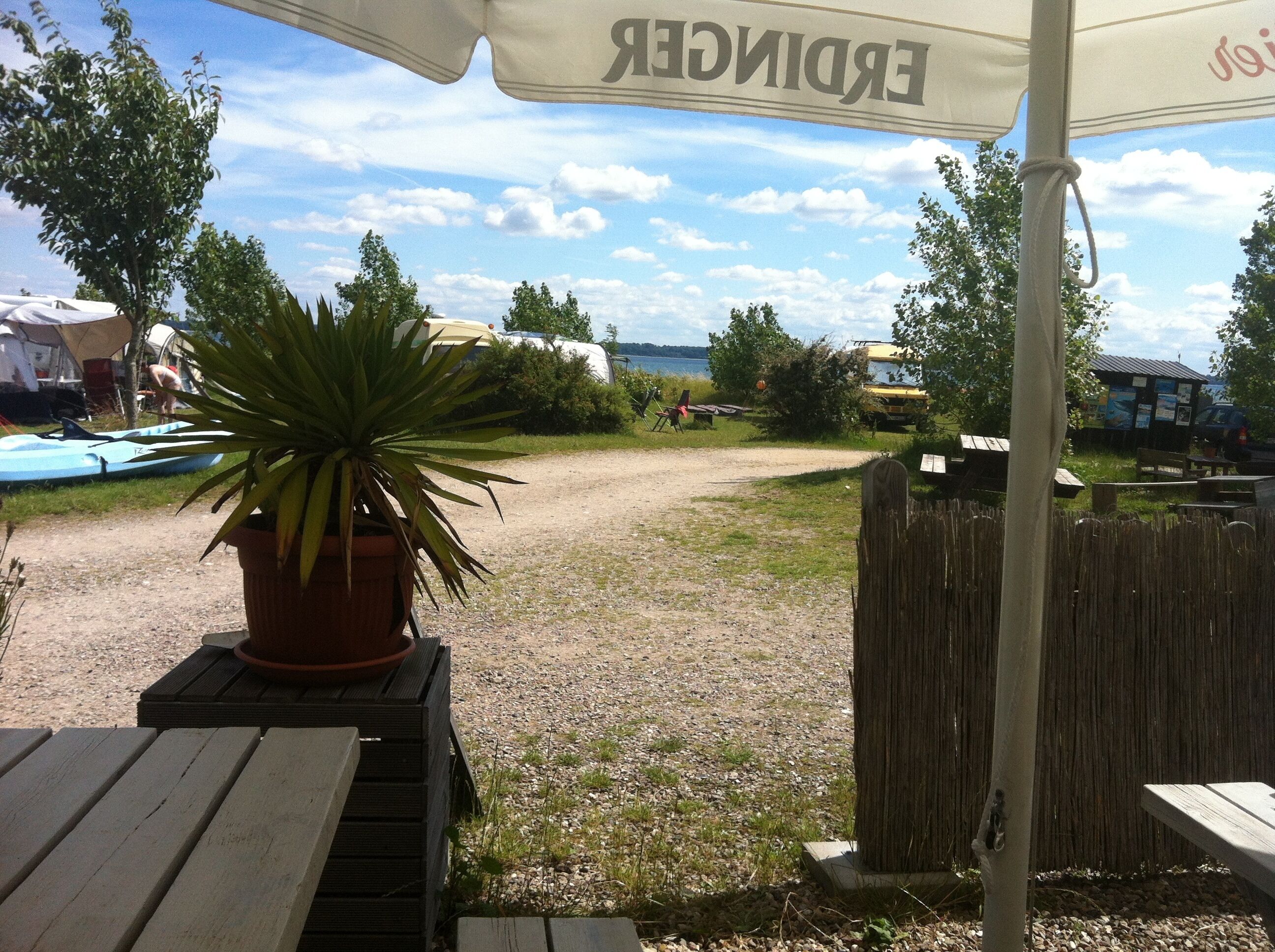Un área de estar al aire libre con una mesa de madera y una planta en maceta, un cercado de madera y vistas a un lago con barcos al fondo.