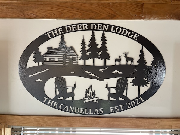 A logo for The Deer Den Lodge, established in 2021, featuring a cabin, trees, deer, and a campfire.