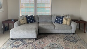 Una sala de estar con un sofá seccional gris, cojines decorativos estampados, un tapete con diseño y dos ventanas con persianas.