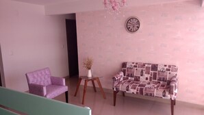 Una sala de estar con un sillón morado, un sofá estampado, una mesita con un florero y un reloj de pared.