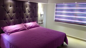 Un dormitorio moderno con un cabecero capitonado, una cama grande con ropa de cama morada, una mesita de noche y un televisor montado en la pared.
