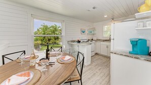 Une cuisine moderne avec des meubles blancs, un coin repas doté d’une table en bois préparée pour deux personnes, et une vue sur les palmiers à travers une grande fenêtre.
