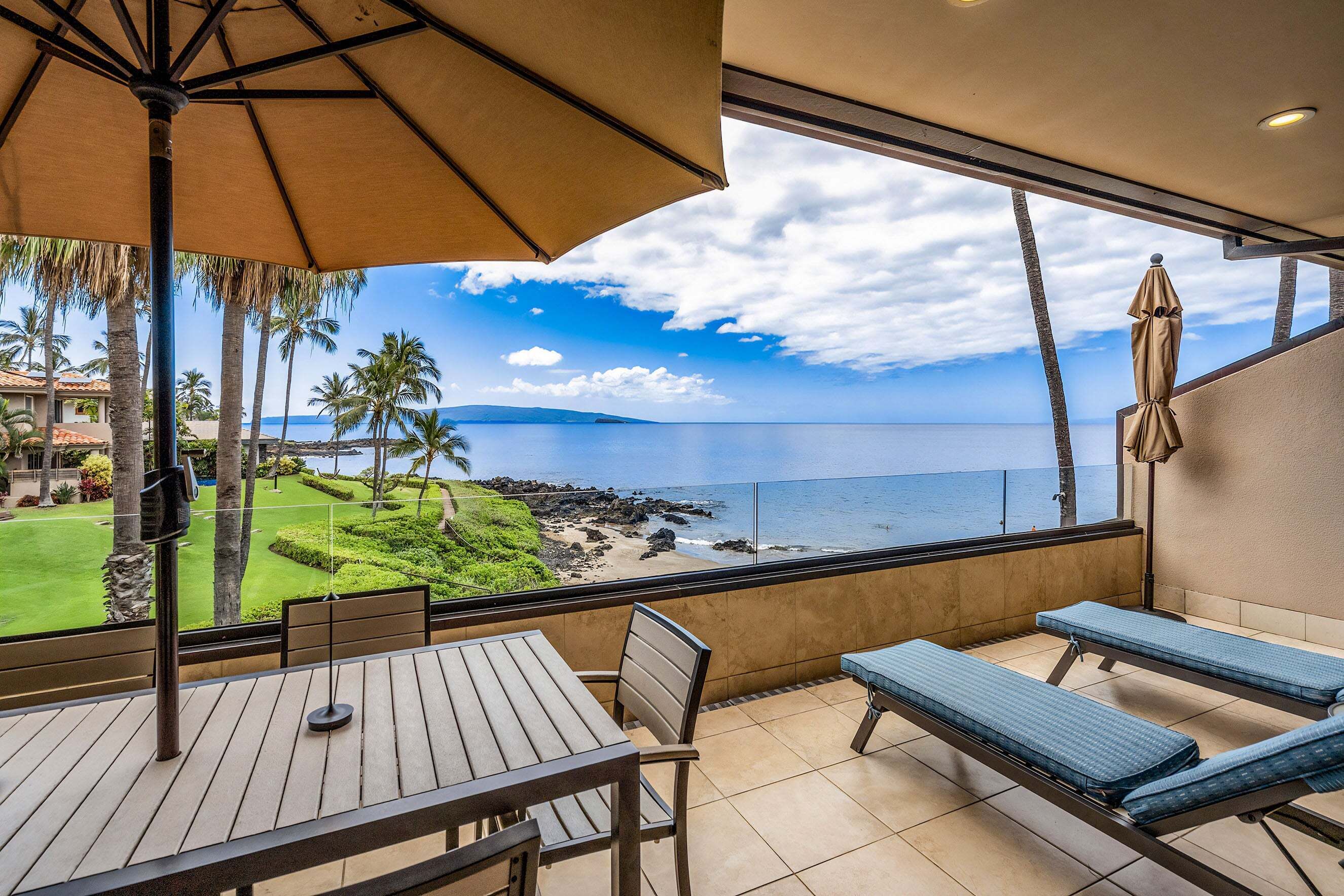 A balcony with a table and chairs, a lounge chair, and a view of the ocean and palm trees.