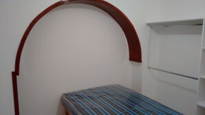 A small, narrow room with a single bed, a white wall, and a red archway.