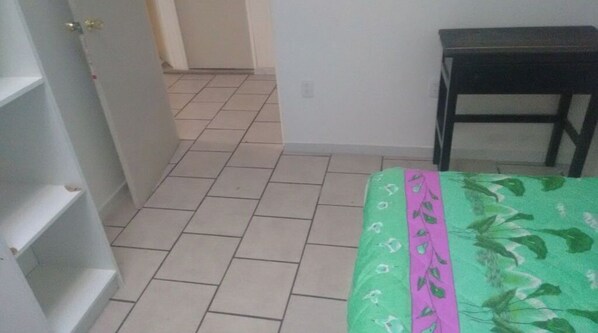Un couloir étroit au sol carrelé, un lit recouvert d’une courtepointe à motifs floraux verts et une petite table en bois.