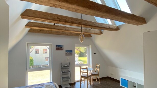 A room with a sloped ceiling, wooden beams, a bed with a striped blanket, a table with chairs, and a view through a window.