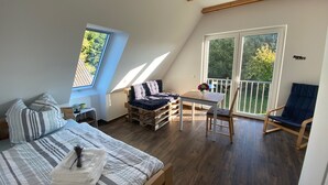 A cozy loft bedroom with a bed, a small table, and a chair. There is a balcony with a chair and a table, and a large window providing a view of the outdoors.