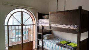 Ein Schlafraum mit Etagenbetten, einem großen Bogenfenster und einer Leiter, die zum oberen Bett führt. Blick auf Gebäude vor dem Fenster.