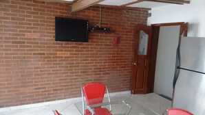 Ein Zimmer mit Backsteinwand, einem angebrachten Fernseher, einem roten Sessel, einem Glastisch und einem Kühlschrank.