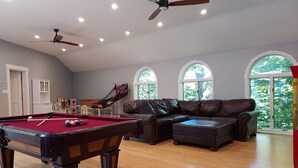 A spacious living room with a pool table, leather sofa, and large windows offering a view of greenery.
