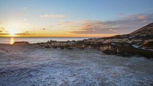 Paesaggio costiero con un tramonto sull'acqua, dune erbose e cielo sereno.