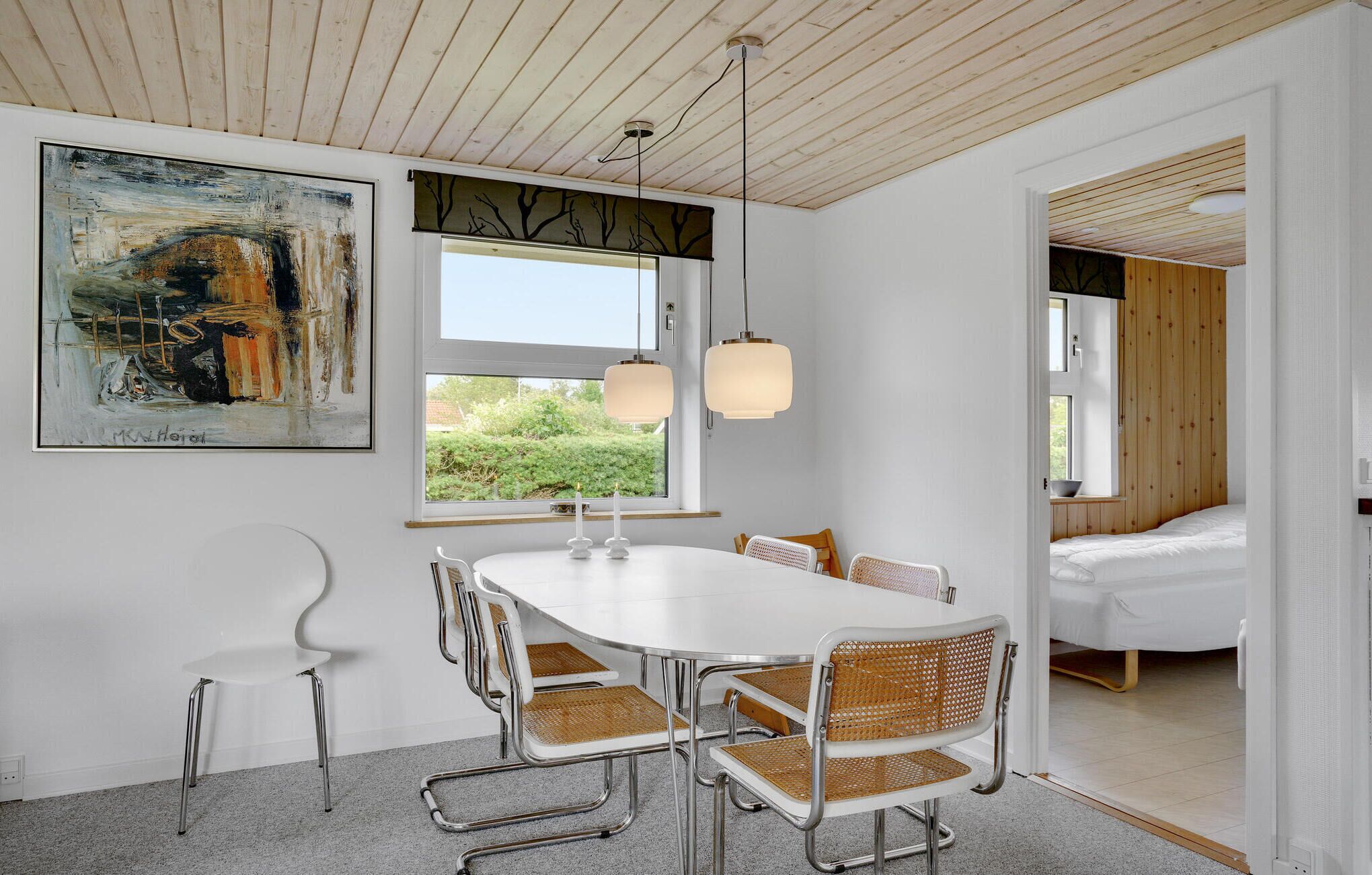 Un espace de salle à manger moderne avec une table et des chaises blanches, une grande peinture abstraite et une vue sur la verdure à l’extérieur.