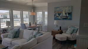 A living room with a white sofa, two armchairs, a dining table, and a painting of sailboats. - Coastal Condo w/ Ocean & Sound Views & Balcony (Emerald Isle)