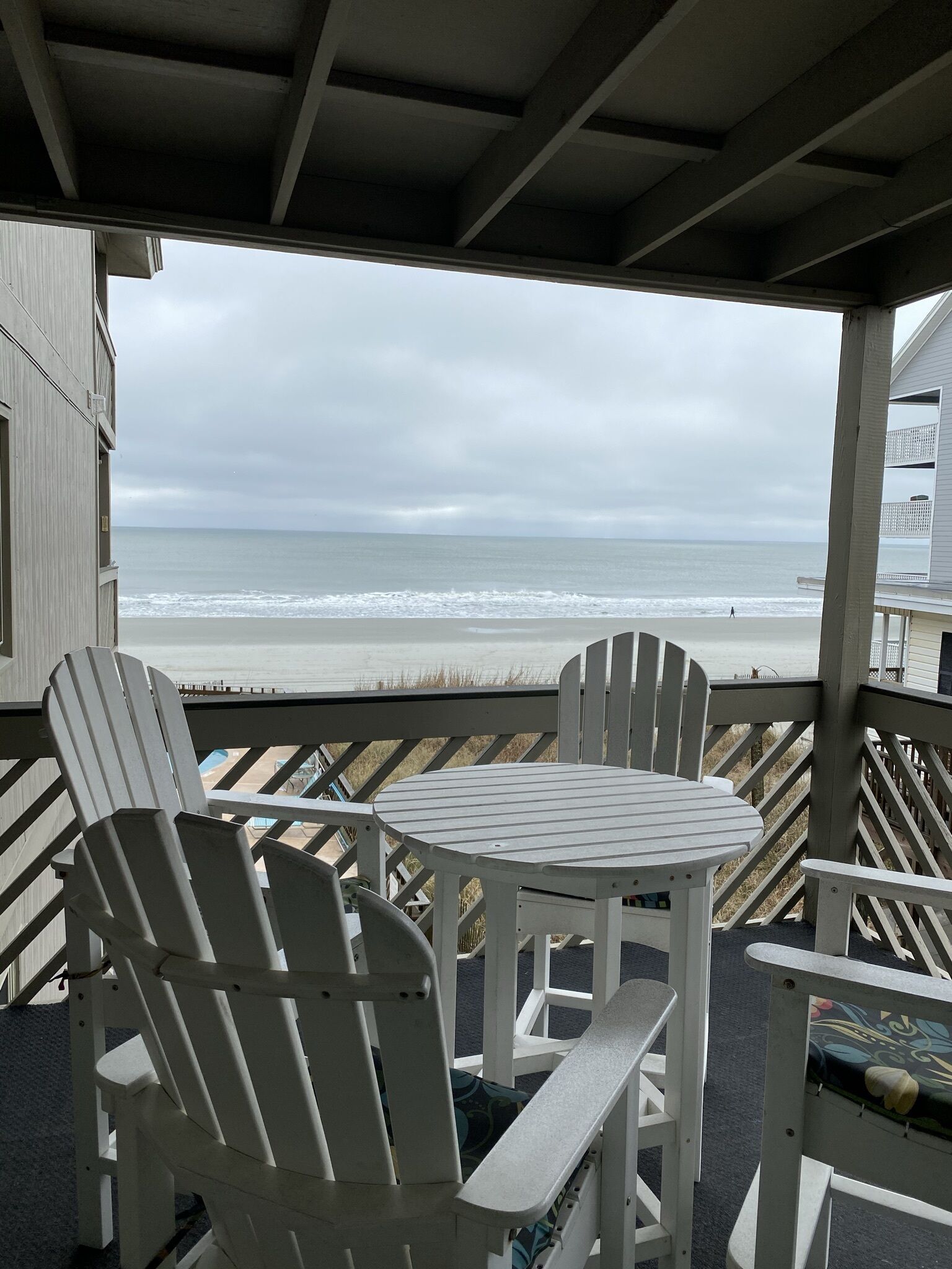 A covered outdoor seating area with Adirondack chairs and a small table, overlooking a beach with waves and a cloudy sky.