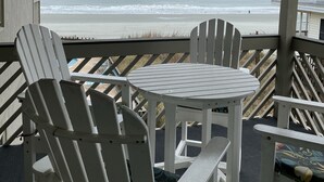 Un espace extérieur aménagé avec une zone de repos couverte, comprenant des fauteuils Adirondack et une petite table, offrant une vue sur une plage avec des vagues et un ciel nuageux.