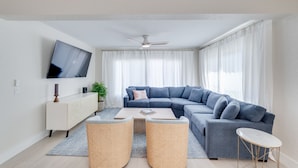 A modern living room with a sectional sofa, a flat-screen TV, a ceiling fan, and a small round table. - NEW REMODEL Steps from private beach access! 4 BR Family Friendly CLEAN  (Fort Myers Beach)