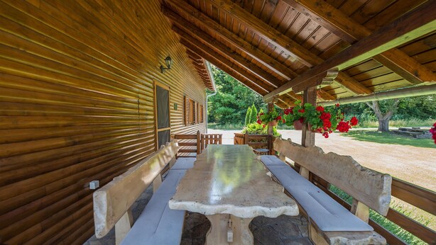 A covered wooden deck with a stone table and benches, surrounded by a garden with red flowers.