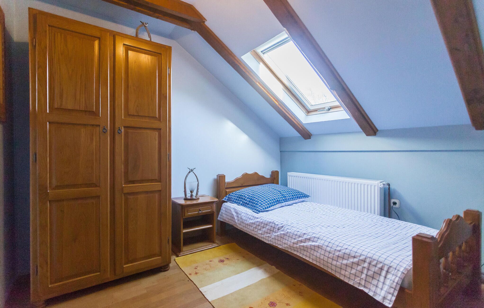 A bedroom with a sloped ceiling, a skylight, a wooden wardrobe, a bedside table with a lamp, a bed with a checkered bedspread, and a radiator.