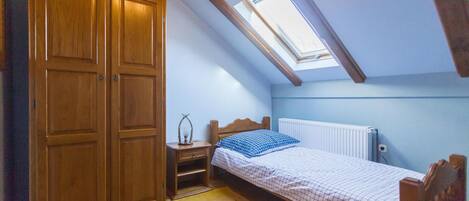 A bedroom with a sloped ceiling, a skylight, a wooden wardrobe, a bedside table with a lamp, a bed with a checkered bedspread, and a radiator.