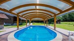 A covered swimming pool with a wooden pergola overhead, surrounded by a grassy area and a wooden building.