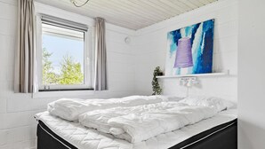 Ett sovrum med en säng, ett fönster med gardiner, en tavla på väggen och en liten växt på en hylla.