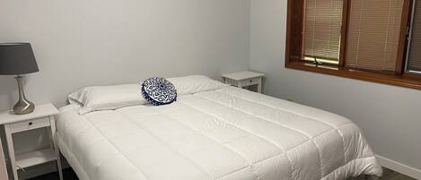 A bedroom with a large bed, a bedside table with a lamp, a small nightstand, and a window with blinds.