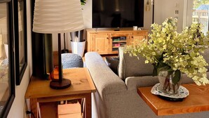 Een moderne woonkamer met een flatscreen televisie, een houten bijzettafeltje, een grijze bank en een kamerplant.