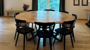 Een eetgedeelte met een ronde houten tafel, zwarte stoelen, een hangende kroonluchter, een groot raam met gordijnen, en ingelijste botanische prints aan de muur.
