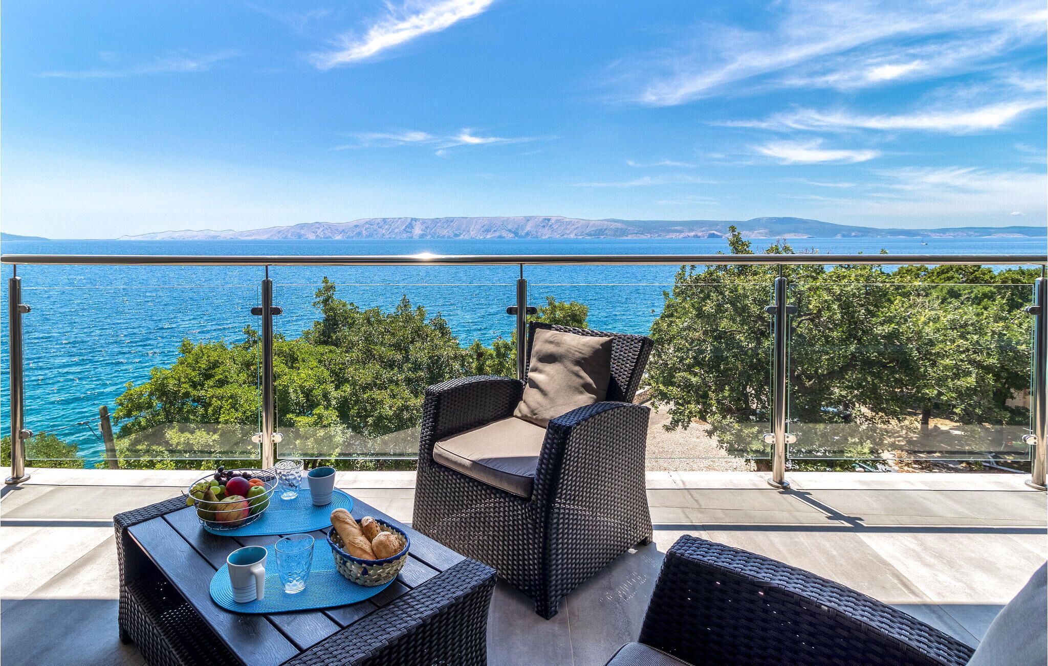 Ein Balkon mit Korbmöbeln, einem Tisch mit Obstschale und Gläsern, und Blick auf das Meer und die Berge.