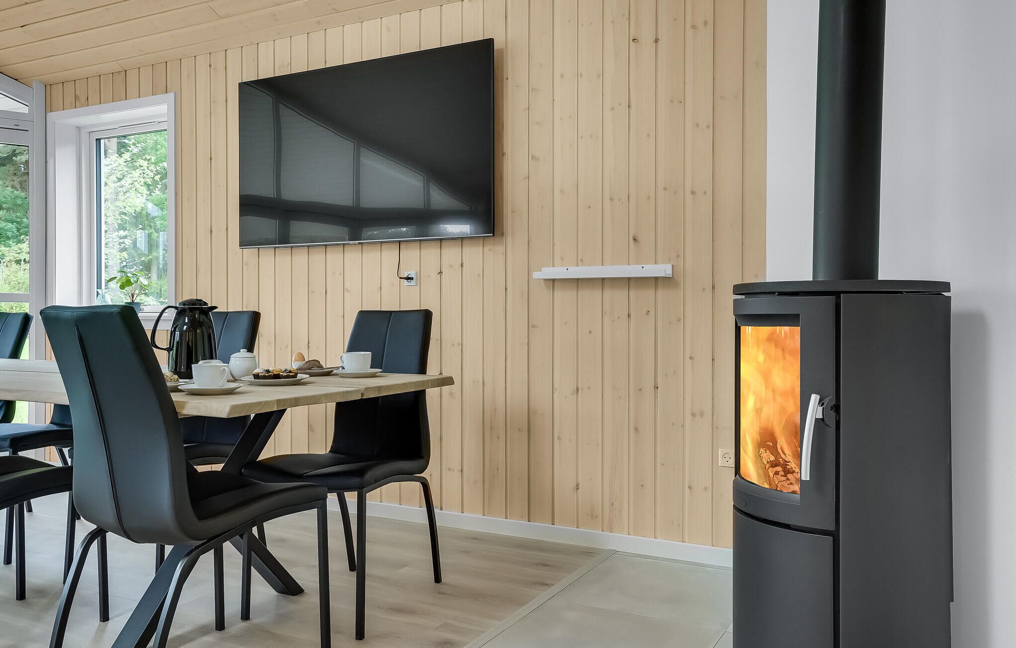 A modern room with a dining table, chairs, a TV mounted on the wall, and a wood-burning stove.