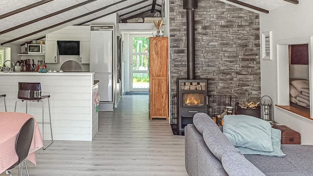 Cocina moderna con armarios blancos, un microondas y un refrigerador. Hay un comedor con una mesa y sillas rosas, y una sala de estar con un sofá gris y una chimenea.