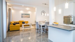 Een moderne woonkamer met een gele bank, een eethoek met een witte tafel, en een keuken met een roestvrijstalen oven.