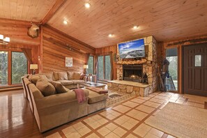 A cozy log cabin interior with a stone fireplace, a large sectional sofa, and a television mounted on the wall.