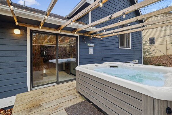 A hot tub outside a house with a wooden deck and a glass door leading to a bathroom.