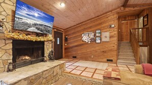 A cozy log cabin interior with a stone fireplace, a large flat-screen TV mounted above it, and a wooden staircase leading to an upper level. - COZY MOUNTAIN ESCAPE! .5mi frm slopes 50ft to trails. HOT TUB! (Beech Mountain)