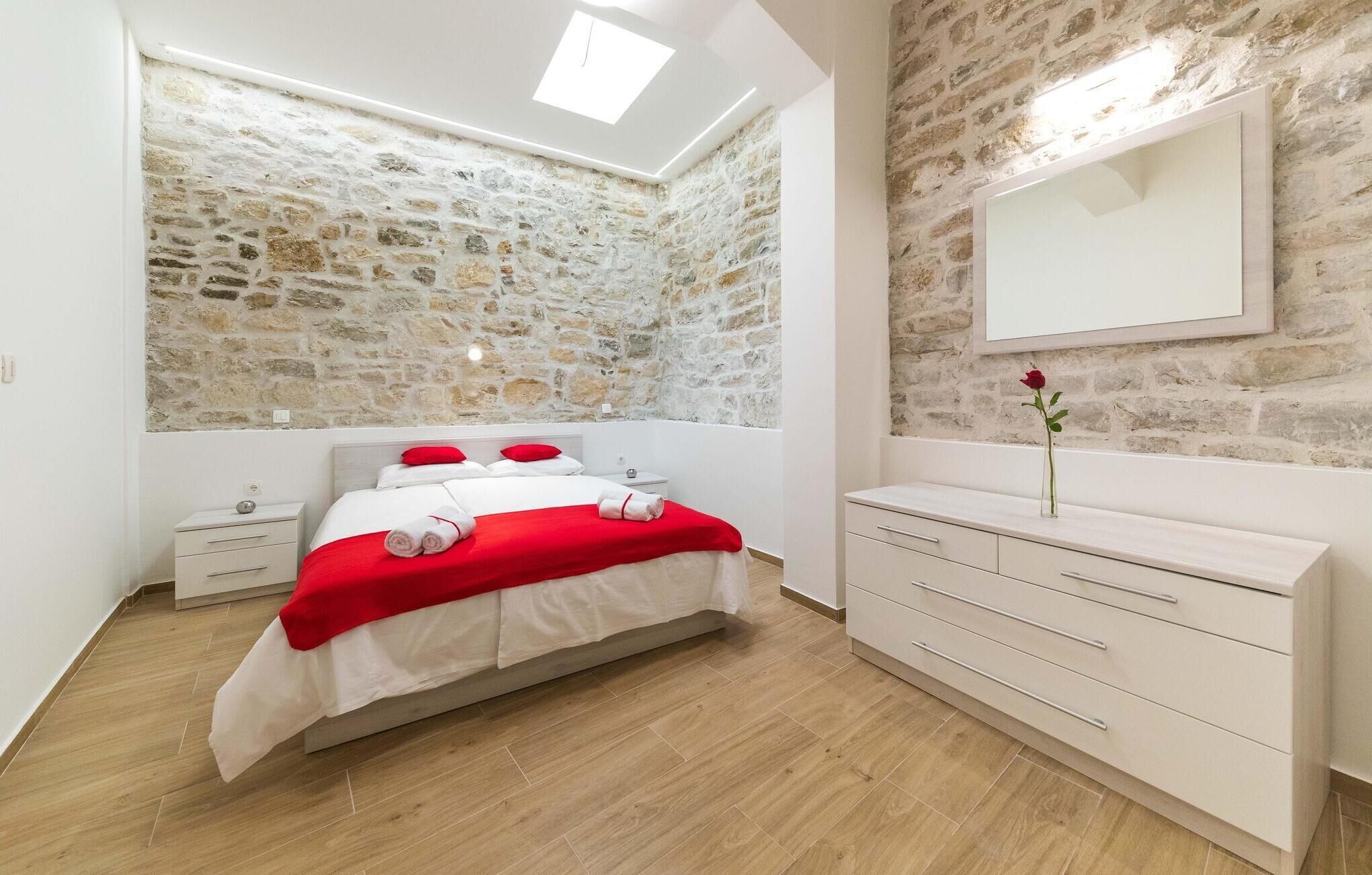 A bedroom with a stone wall, a bed with red and white bedding, a white dresser, and a mirror.
